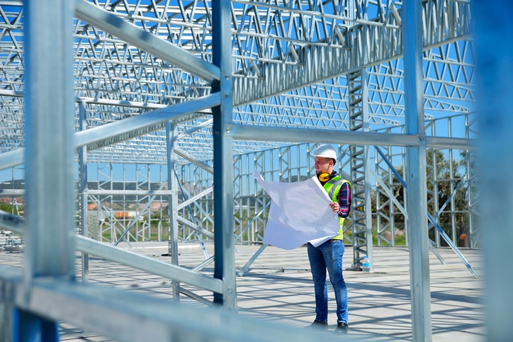 A construction worker wearing PPE evaluating the steel structure with a blueprint in their hands.