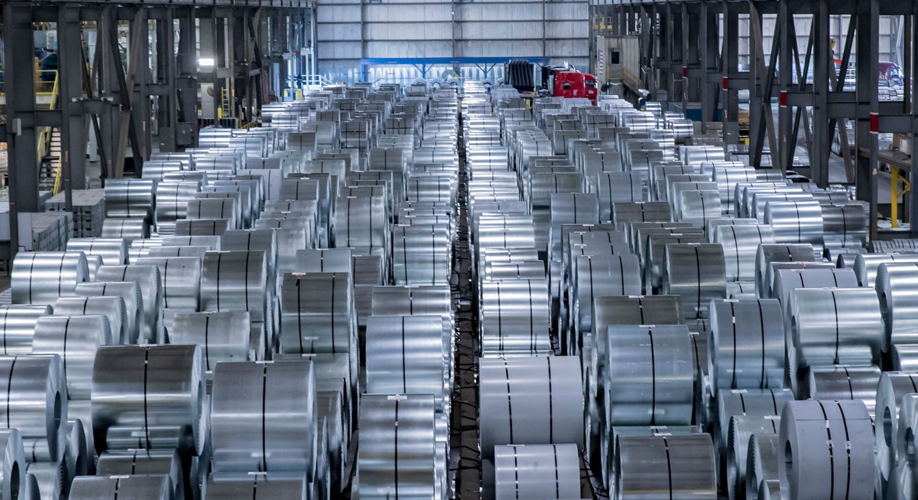 Inside a warehouse, hundreds of stacked steel coils sit held together with black bands.