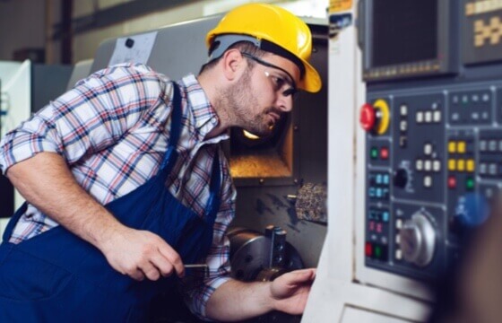 Safety employee holding a screwdriver inspecting piece of machinery while wearing protective gear.